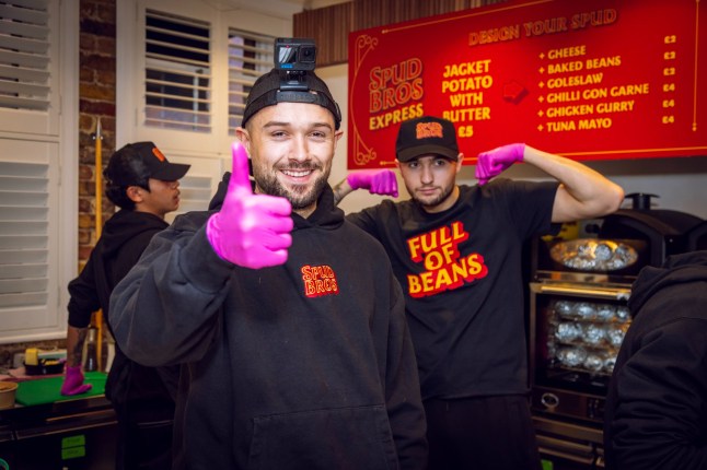 Spud Bros, Jacob Nelson (left) and Harley Nelson stood in a kitchen making jacket potatoes. They are wearing their own merch and pink gloves.